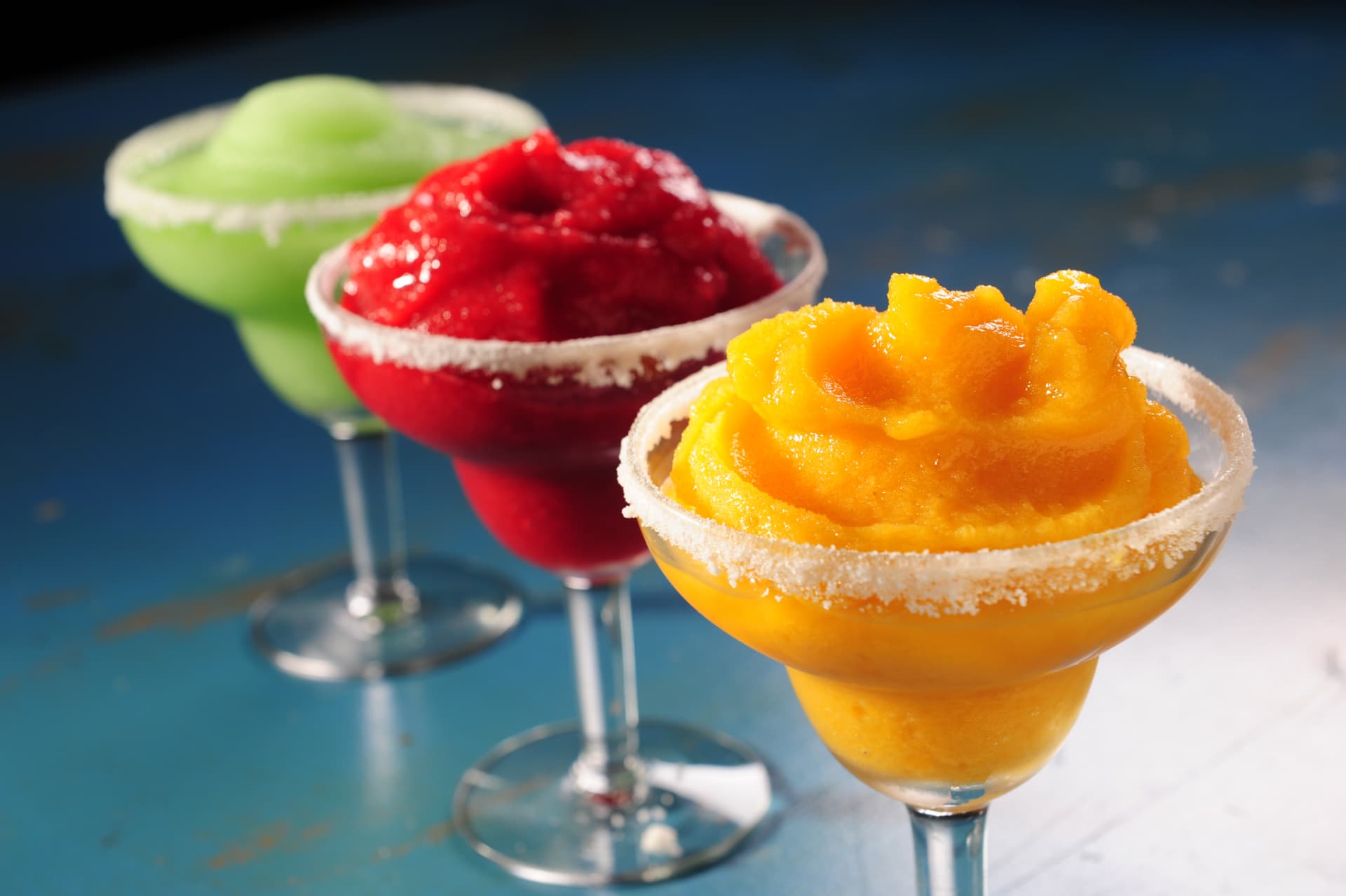 Three colorful frozen margaritas in sugar-rimmed glasses